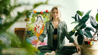 A woman in a green leather jacket sits on the floor surrounded by plants, taking a moment to relax