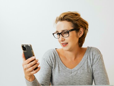 A woman wearing a gray long-sleeve shirt takes a selfie and keeps checking her phone