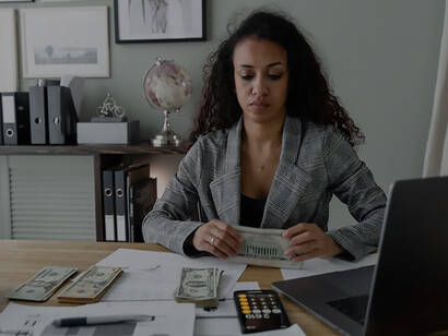 In her office, an African woman engages in thoughtful financial planning, surrounded by both traditional coins and modern computer technology