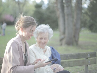 An elderly woman smiling as her younger relative is using a piece of technology 