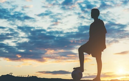 A young footballer silhouetted against the evening sky with a ball at their feet, representing dreams that reach far beyond the horizon