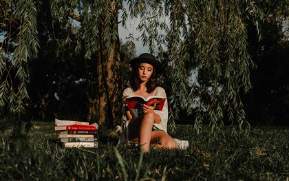 Jovem lê um livro sob a sombra de uma árvore, cercada por outros livros. A imagem remete ao início de uma busca interior que une contemplação, conhecimento e silêncio