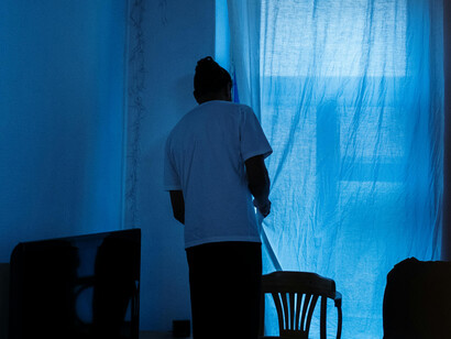 Man in a white T-shirt standing alone near a sliding door at home in a calm, blue-lit apartment