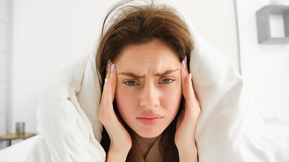 A woman lies in bed under white sheets, frowning in pain from menstrual migraines in the morning