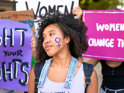 A protester standing resolute with placards rising behind her, symbolising the courage fuelling the demand for gender equality