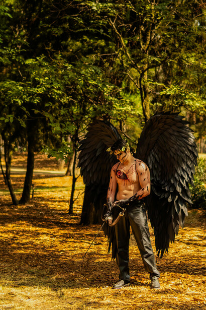 A shirtless man posed in an angel costume with a bow, his wings creating a striking fallen-angel aesthetic and symbolic imagery