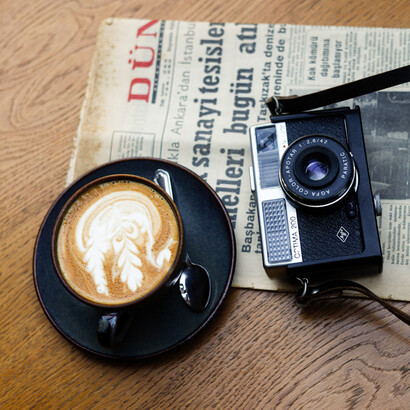 Un periódico, una cámara y una taza de café se encuentran sobre una mesa, como una pequeña escena del oficio periodístico: informarse, mirar y contar
