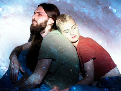 Una coppia è seduta, con la donna appoggiata sulla spalla dell'uomo, entrambi illuminati dalla proiezione di una galassia che avvolge lo sfondo