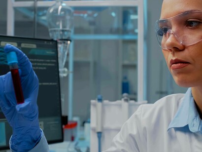 Primo piano di una scienziata in camice bianco e occhiali protettivi che tiene in mano una provetta contenente un campione di liquido, analizzandola di fronte a un monitor di laboratorio