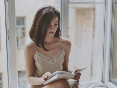 A woman in a nightdress sits on a windowsill, reading a book and drinking coffee