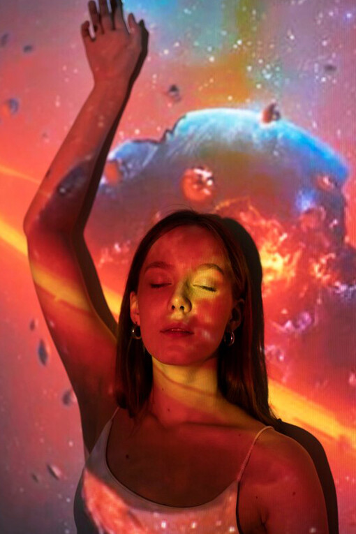 A young woman standing beneath a celestial projection, blending into a world of surreal landscapes, cosmic energy, and infinite dreamlike spaces