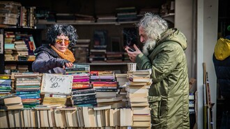 Uma mulher procura um livro; ao seu lado, um homem consulta o celular, ambos cercados por centenas de exemplares. A cena coloca frente a frente duas formas de acesso ao conhecimento e sugere que o desafio do leitor contemporâneo não está apenas em escolher entre o papel e a tela, mas em aprender a transitar entre diferentes possibilidades de leitura. Papel e tecnologia convivem na mesma cena, lembrando que, embora os suportes se transformem, permanece o desejo de ler, descobrir e alcançar novos conhecimentos