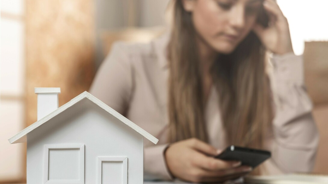 A woman holds her phone while standing behind a miniature wooden house, deep in thought