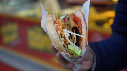 A man holding a doner kebab in front of a bustling street food restaurant