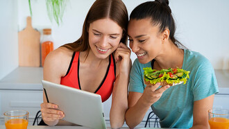 A woman in a kitchen, surrounded by healthy food, examines an online information source on the table