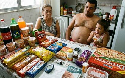 Uma família diante de uma mesa repleta de produtos ultraprocessados, em uma cena que traduz o contraste entre a abundância de alimentos industrializados e o acesso cada vez mais difícil à comida de verdade. Quando os produtos ultraprocessados ocupam a mesa com facilidade, comer comida de verdade pode deixar de ser uma escolha cotidiana e se transformar em privilégio