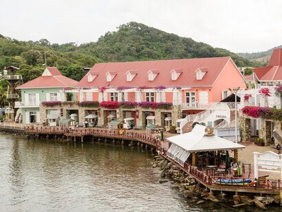 The building features a distinctive red roof, pink exterior walls, and a stone-based arcade with shops at the waterfront level