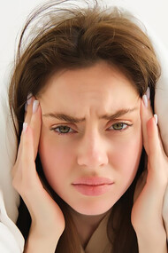 A woman lies in bed under white sheets, frowning in pain from menstrual migraines in the morning