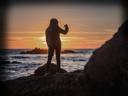 A woman stands before a sunset on the sea, taking a picture