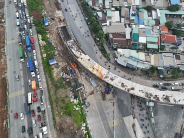 Vehicles trapped beside a massive unfinished infrastructure project, representing the gap between ambitious development and everyday public realities