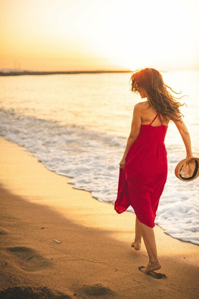 De costas, uma mulher de vestido vermelho e chapéu de palha caminha pela praia, junto ao mar. Afastar-se por alguns instantes do ruído cotidiano pode abrir espaço para escutar a si mesmo e reconhecer o caminho que realmente faz sentido seguir