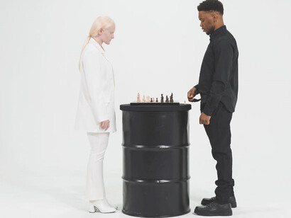 Amidst a captivating backdrop, a woman clad in a pristine white suit and a man in a suave black suit engage in a game of chess atop sleek black barrels, their strategic minds at play