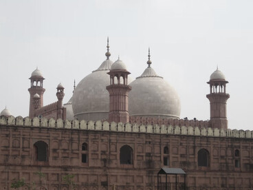 The Badshahi Mosque, glowing in the morning light, stands tall in red and white hues, seen from Minar-e-Pakistan and framed by lush trees