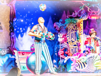 A colorful juggler on the fantasy stage as another performer nearby plays the violin to his act