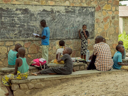 Young students learning beside a weathered chalkboard, symbolising both the resilience of learners and the urgent need for better educational infrastructure