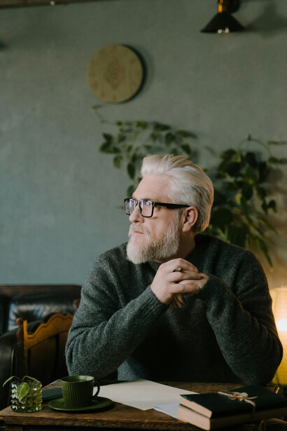 An elderly man, immersed in contemplation, writes at a table with a pen in hand