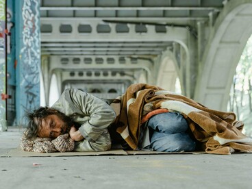 Um homem de jaqueta verde está deitado sob um viaduto urbano, envolto em uma manta. A imagem remete à luta diária pela sobrevivência e à importância de políticas públicas que assegurem proteção e dignidade às pessoas em situação de rua
