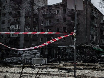 Edifícios danificados, veículos atingidos, destroços espalhados pelo chão e fitas de isolamento compõem a paisagem de uma rua de Kyiv após um ataque. A destruição da infraestrutura urbana revela uma das faces mais persistentes da guerra russo-ucraniana: mesmo longe das posições de combate, os espaços de convivência e a vida cotidiana dos civis permanecem expostos à violência
