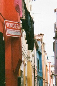 Un vicolo stretto a Burano, Italia, fiancheggiato da edifici dai colori vivaci, con un cartello rosso con la scritta "Vendesi" su uno degli edifici
