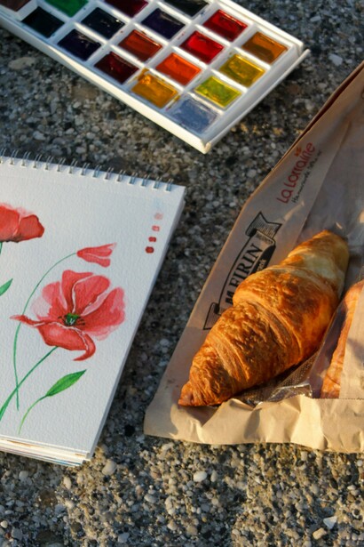 Um bloco de aquarelas, uma caixa de tintas e um croissant compõem um cenário de criação artística ao ar livre. Com cafés, esboços e pequenas pausas, François transforma as impressões da viagem em futuras pinturas