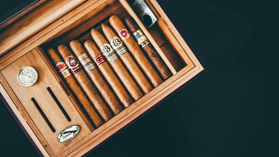 A hand-made wooden cigar box, open to reveal a neatly arranged row of cigars with varied bands, alongside a silver cigar cutter