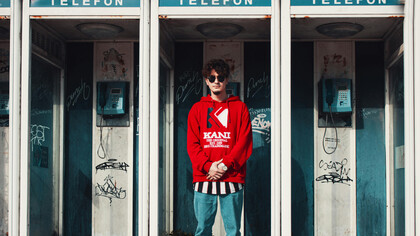 A man in a red hoodie stands inside a phone booth, lost in thought or conversation