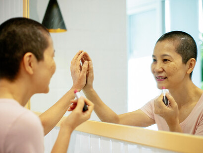 An Asian woman with breast cancer applies lipstick while looking in the mirror