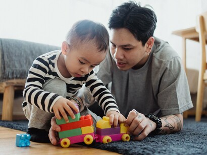 Um pai e seu filho compartilham uma brincadeira com blocos coloridos, explorando formas e possibilidades de construção. Nas interações cotidianas, a criança experimenta o ambiente, observa resultados e desenvolve novas maneiras de compreender o mundo