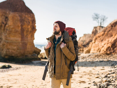 Adventurous young traveler carrying a backpack along an autumn sea shore, dressed warmly in a jacket and hat