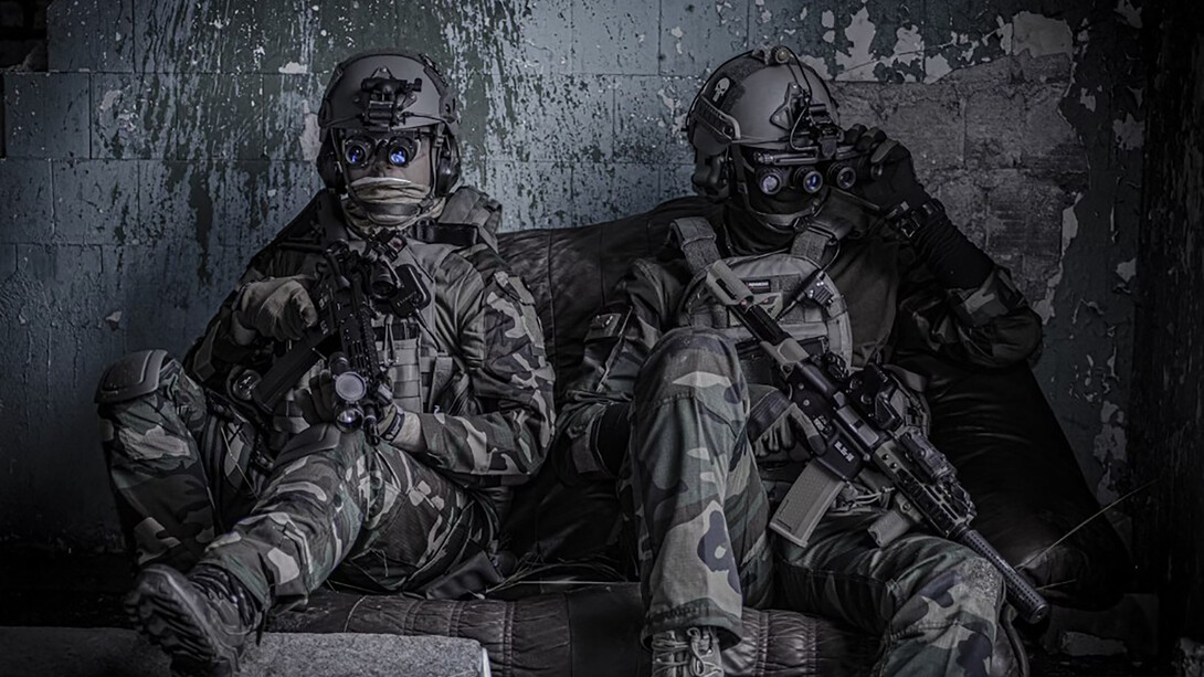 Two officers sit on a the floow, dressed as airsoft players wearing protective glasses, helmets, and holding airsoft guns