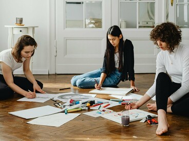 Papéis em branco, rabiscos e cores dividem o espaço com três mulheres em um momento de criação espontânea. Nem tudo precisa ser útil. Às vezes, o processo é o próprio caminho