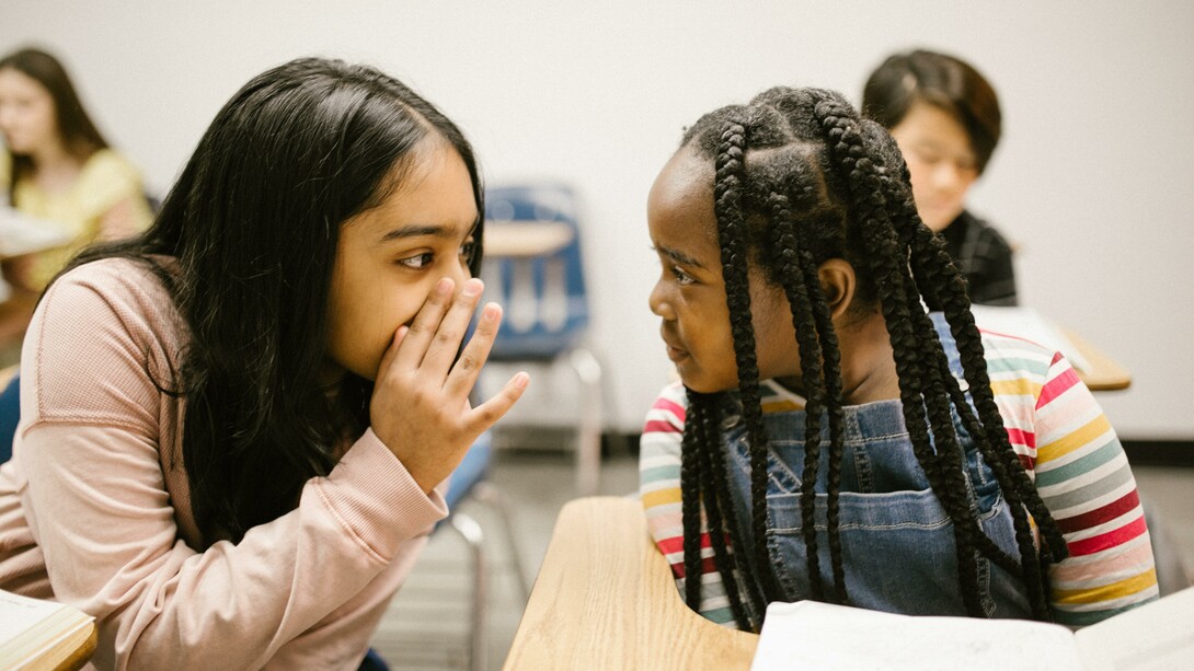 Two girls whispering during a lesson, inside a classroom, at school