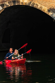 A man and woman paddle a red kayak along a river in Utrecht, the Netherlands, under the bright daytime sky