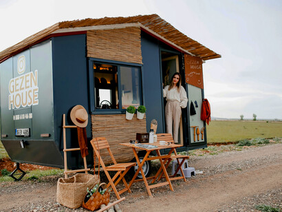 A man and a woman sit on a brown wooden chair near a black tiny portable house