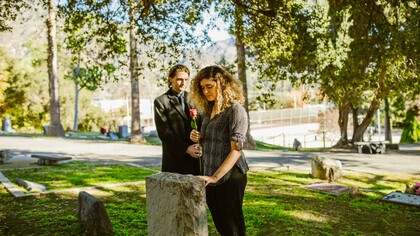 Mulher de camisa preta ao lado de uma lápide de concreto cinza enquanto segura uma flor