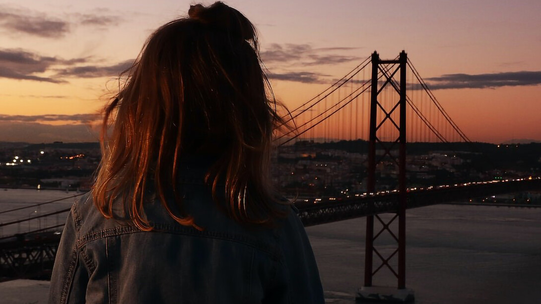 A woman stands gracefully in front of a Lisbon bridge at the enchanting backdrop of a Portuguese sunset