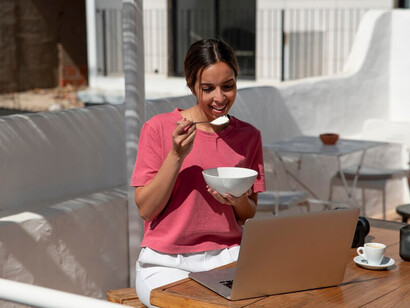 A woman sitting at a terrace table with her laptop, surrounded by work materials, facing away from the camera