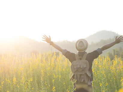Un escursionista si trova in un campo di fiori gialli con le braccia spalancate verso il cielo e il sole che tramonta o sorge sulle colline