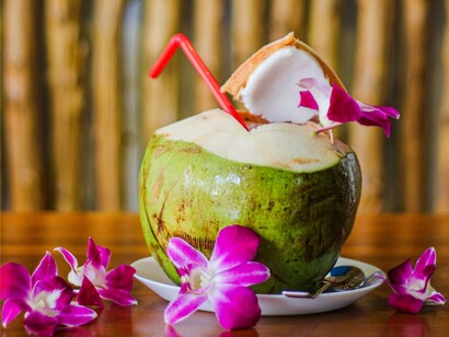 Um coco fresco servido sobre uma mesa de madeira ganha um toque ornamental com vibrantes orquídeas cor-de-rosa. A cena evoca a atmosfera tropical de um destino em que a experiência de viajar também passa pelos sabores, pelas cores e pela proximidade com a natureza