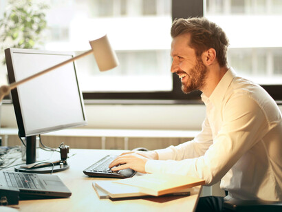 Seated on a black rolling chair, a man in a white dress shirt smiles warmly while working at his black computer setup